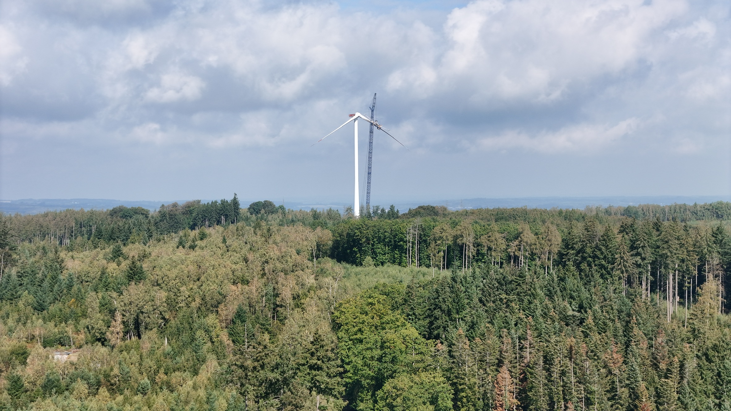 Windrad und Kran über dem Wald unter einem wolkigen Himmel